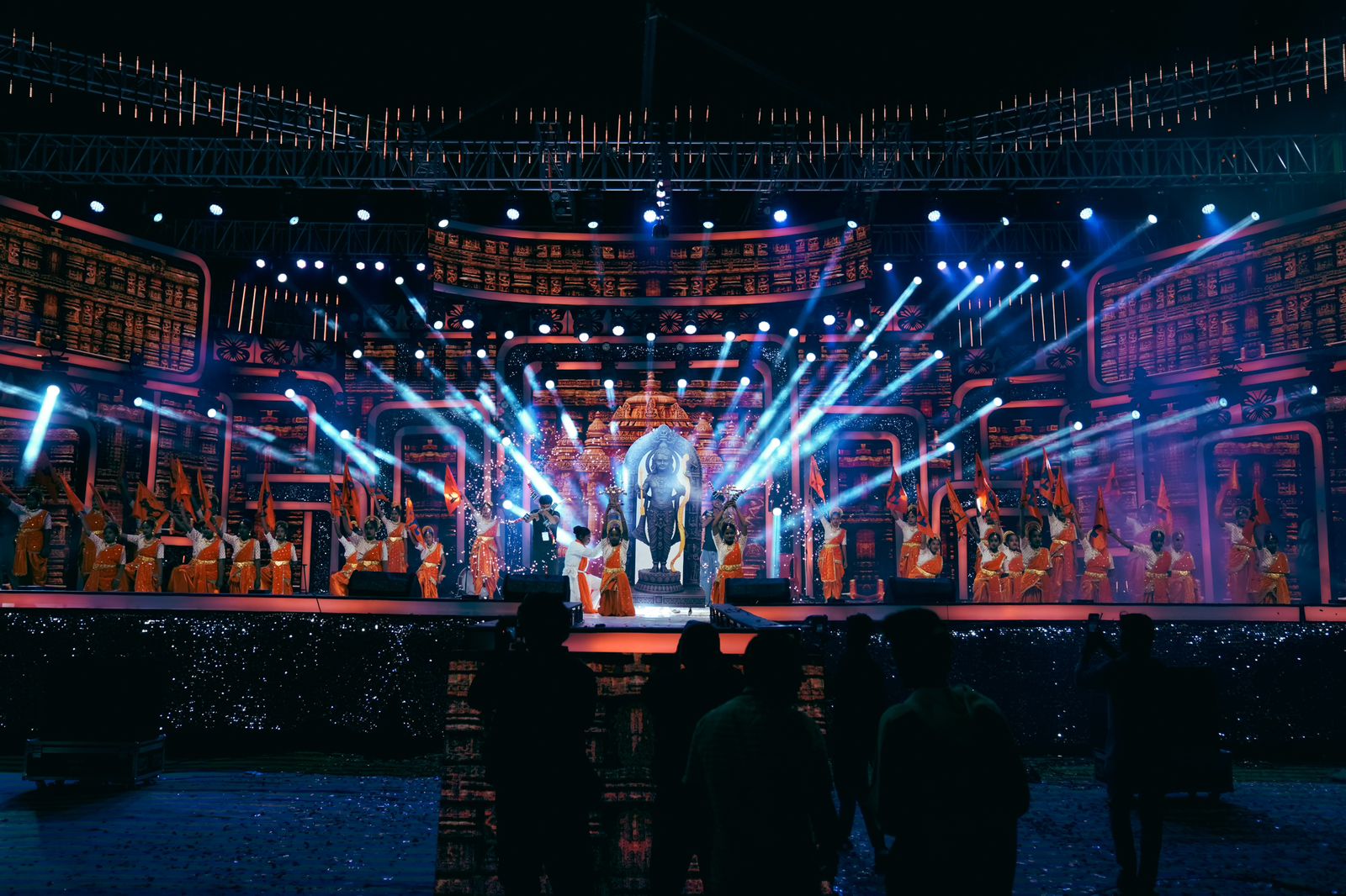 Large-scale cultural event with temple-inspired LED backdrop, beam lighting, and performers with flags in Karnataka